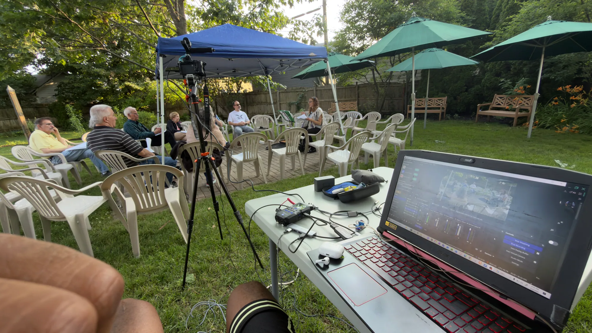 Outdoor livestream control station with a camera on tripod, laptop preview, and production equipment under a canopy.