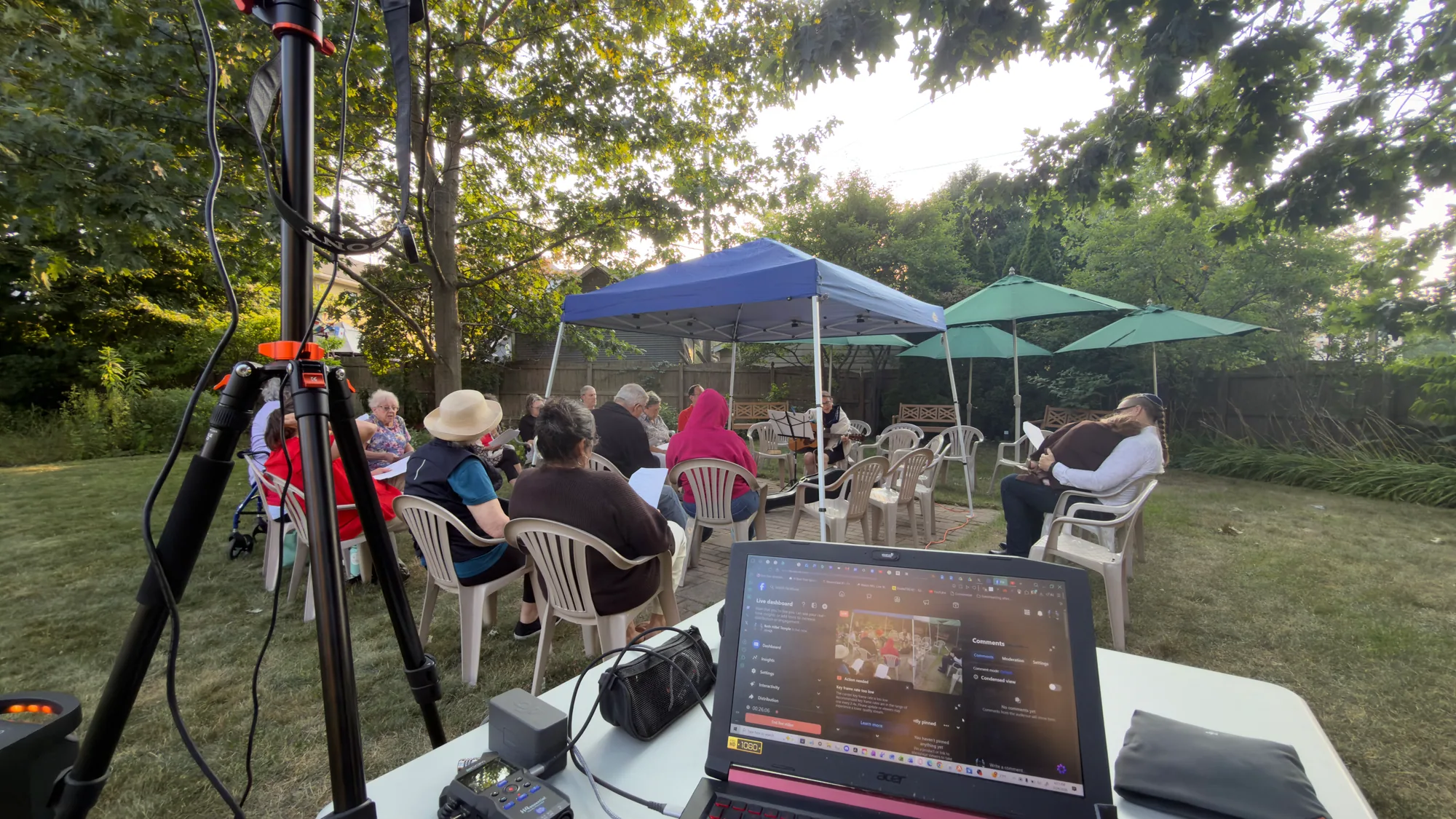 Wide view of an outdoor service gathering with the camera tripod and livestream control table in the foreground.