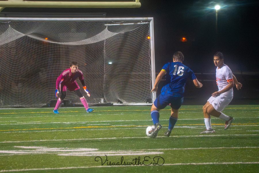 A blue-shirted attacker advances toward a goalkeeper in front of the goal.