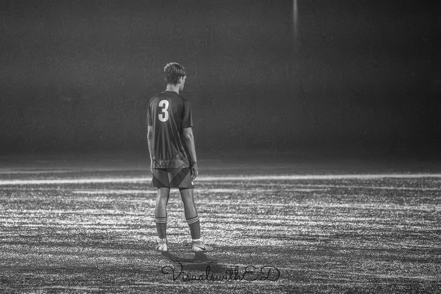 Black-and-white view of a soccer player standing alone on the rain-lit field.
